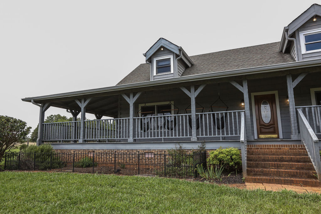 Front view of on-site wedding venue lodging featuring a wraparound porch and stone chimney surrounded by open green space.