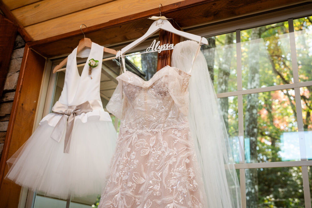 A wedding gown and flower girl dress hang in the window with fall foliage outside