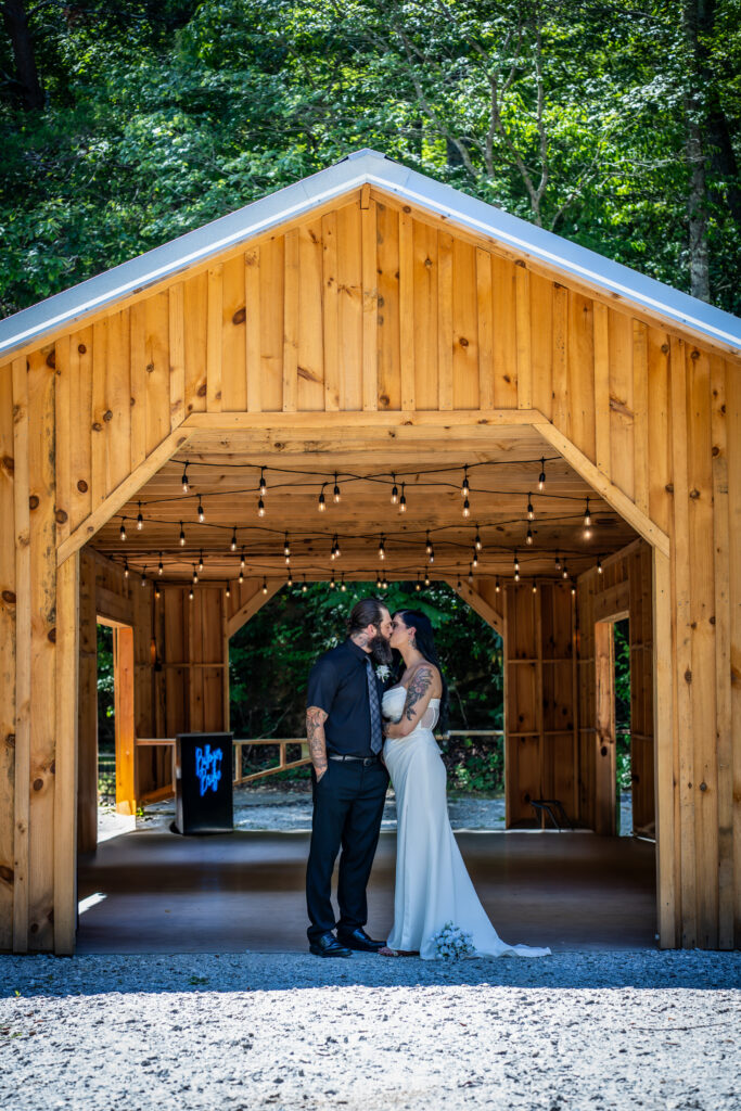 A bride and groom kiss outside a natural wood covered bridge with string lights in the background