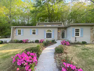 A cozy log cabin nestled in the woods along a river, with blooming azaleas along the walkway
