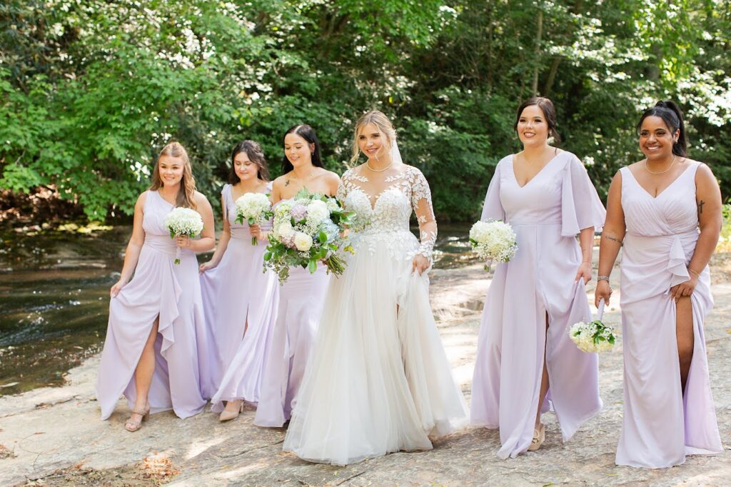 Bridesmaids dressed in lilac complement our beautiful bride in her breathtaking wedding dress as they pose for wedding day photos near the riverfront at our Greenville S wedding venue. 