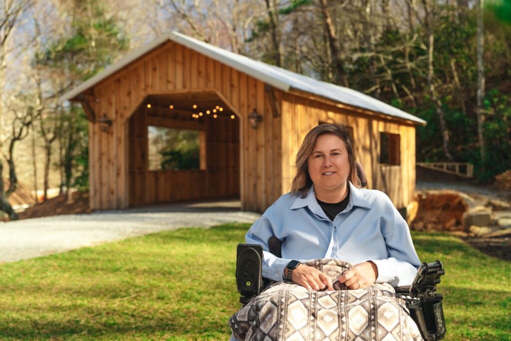 Woman in a wheelchair outdoors in front of a wooden cabin, covered with a patterned blanket, on a sunny day in a yard.
